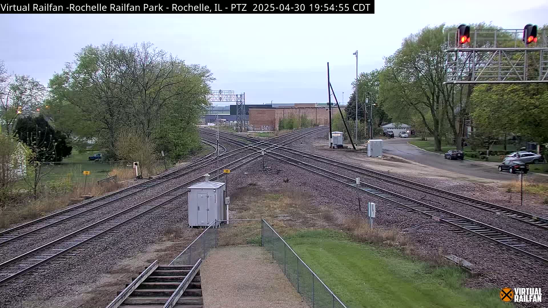 A railway crossing with multiple tracks intersects a road under a partly cloudy sky, surrounded by trees and some buildings in the distance.