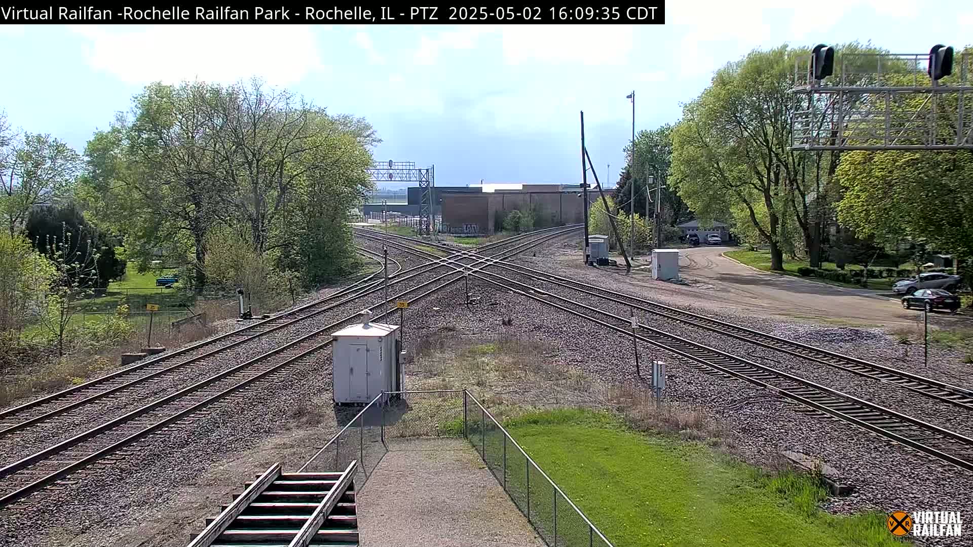 A partly sunny day reveals a railroad crossing with multiple tracks intersecting, surrounded by trees and a small grassy area.