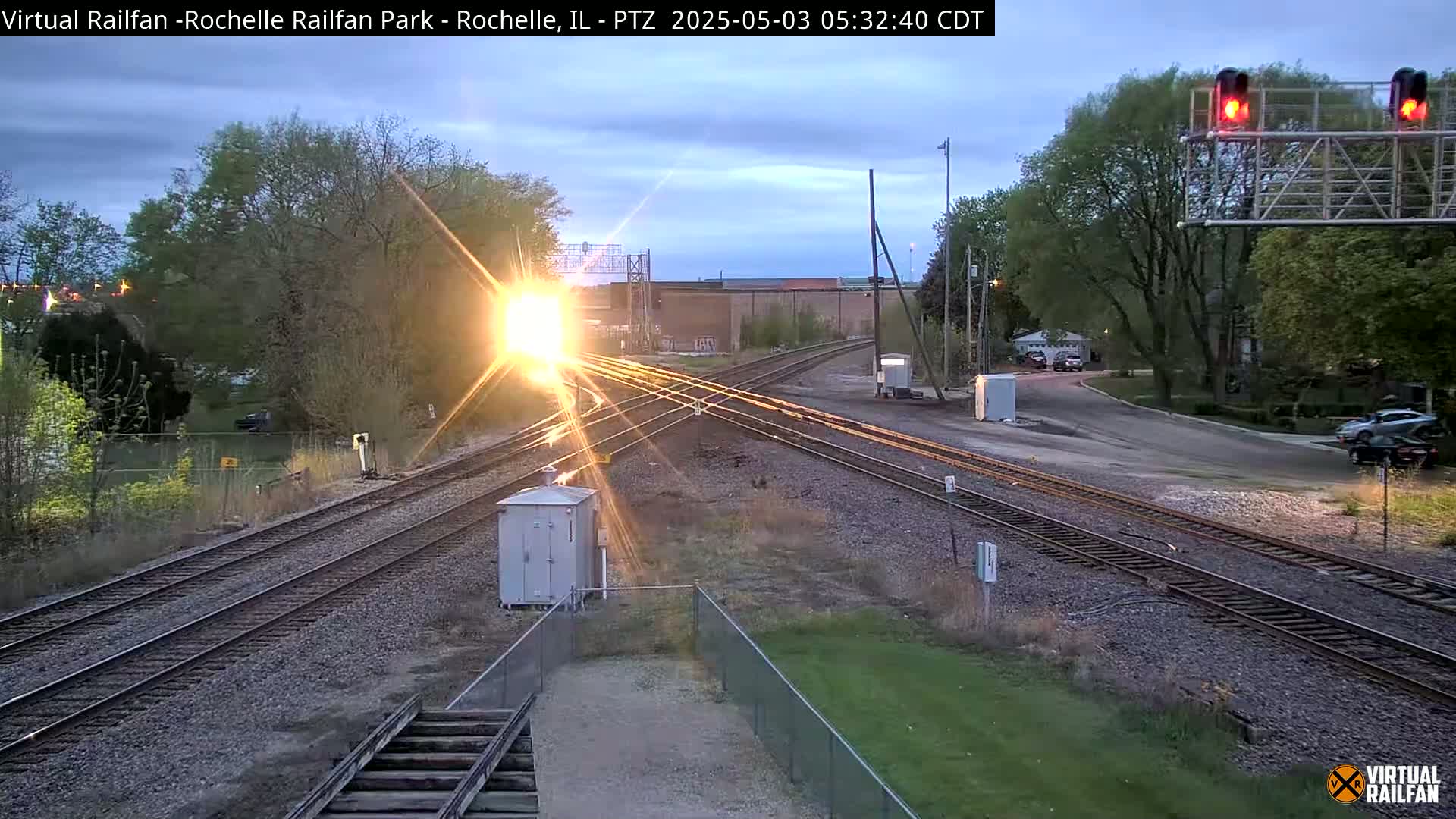A bright sunrise shines over a train track intersection, with trees and buildings visible in the background under a partly cloudy sky.