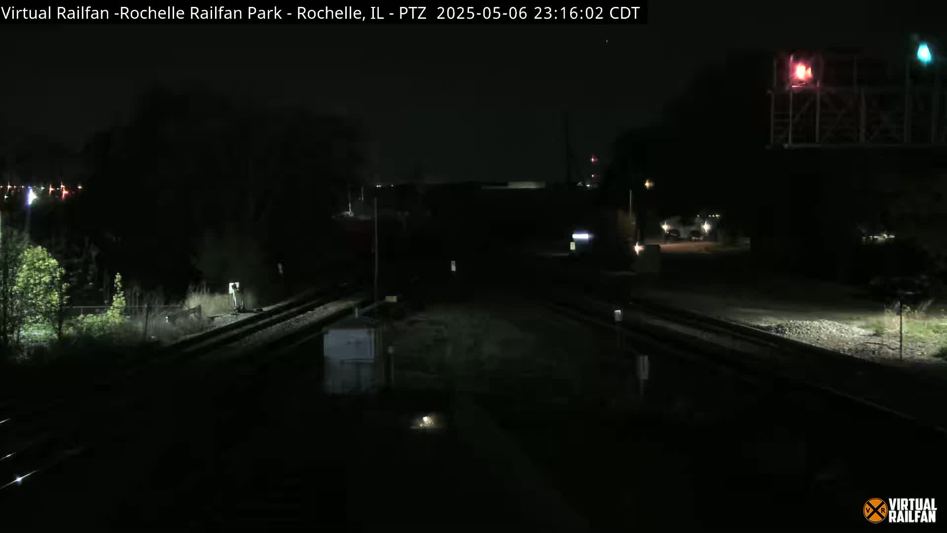 A nighttime view of railroad tracks intersecting under a bridge, with sparse vegetation and a few distant lights visible.