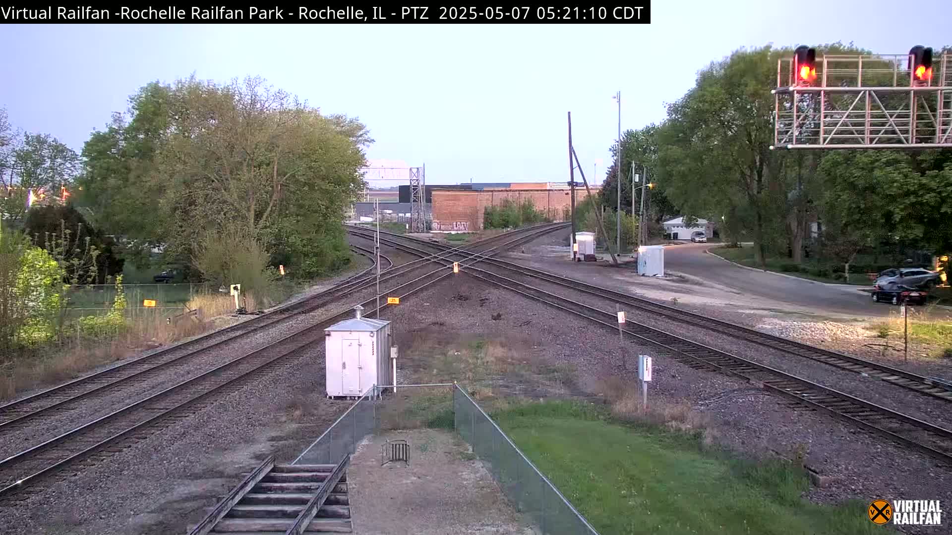 A railroad crossing with multiple tracks under a twilight sky, surrounded by trees and some buildings in the distance.