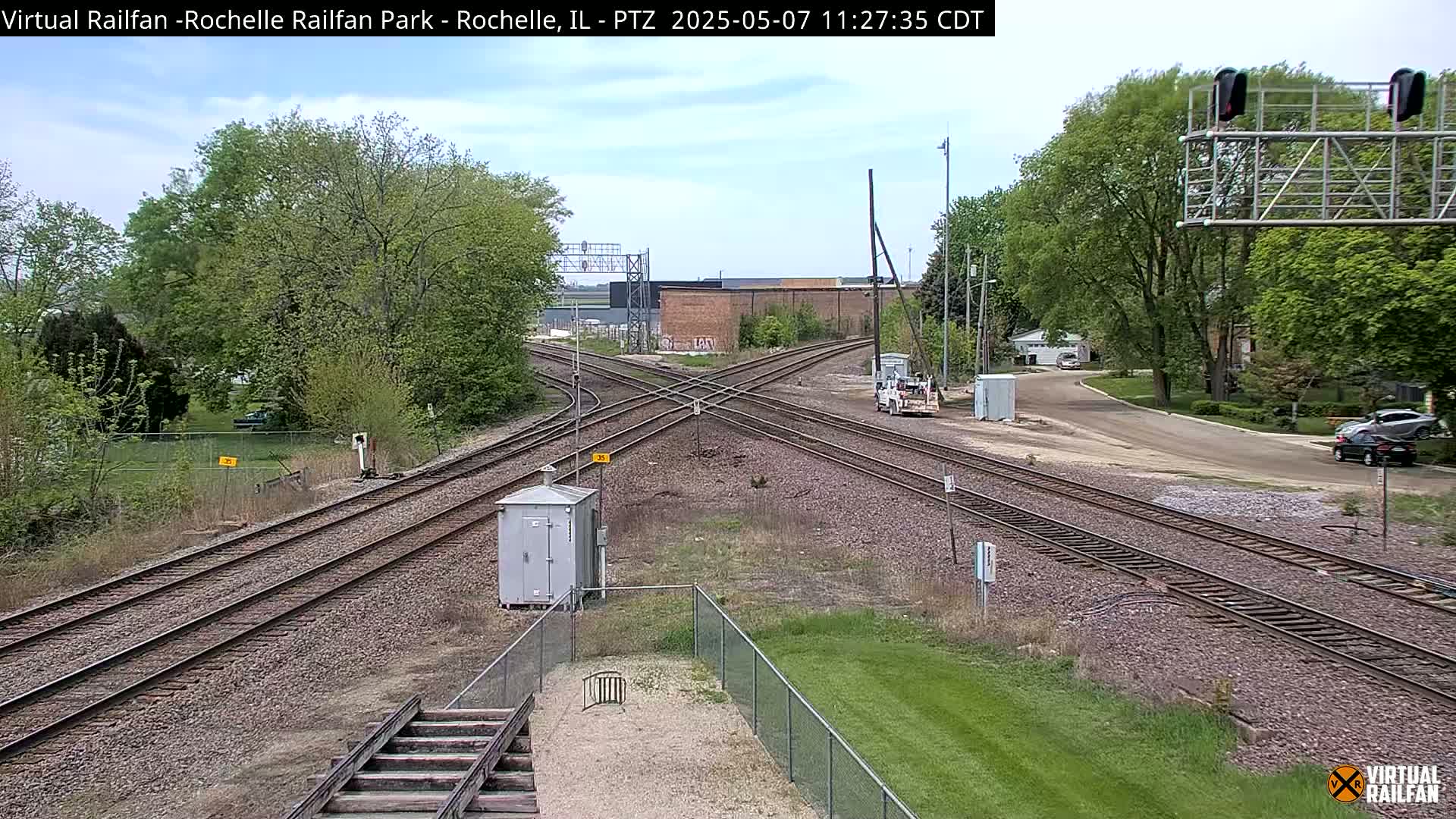 A mostly sunny day shows a railroad crossing with several train tracks intersecting, surrounded by lush green trees and grass.
