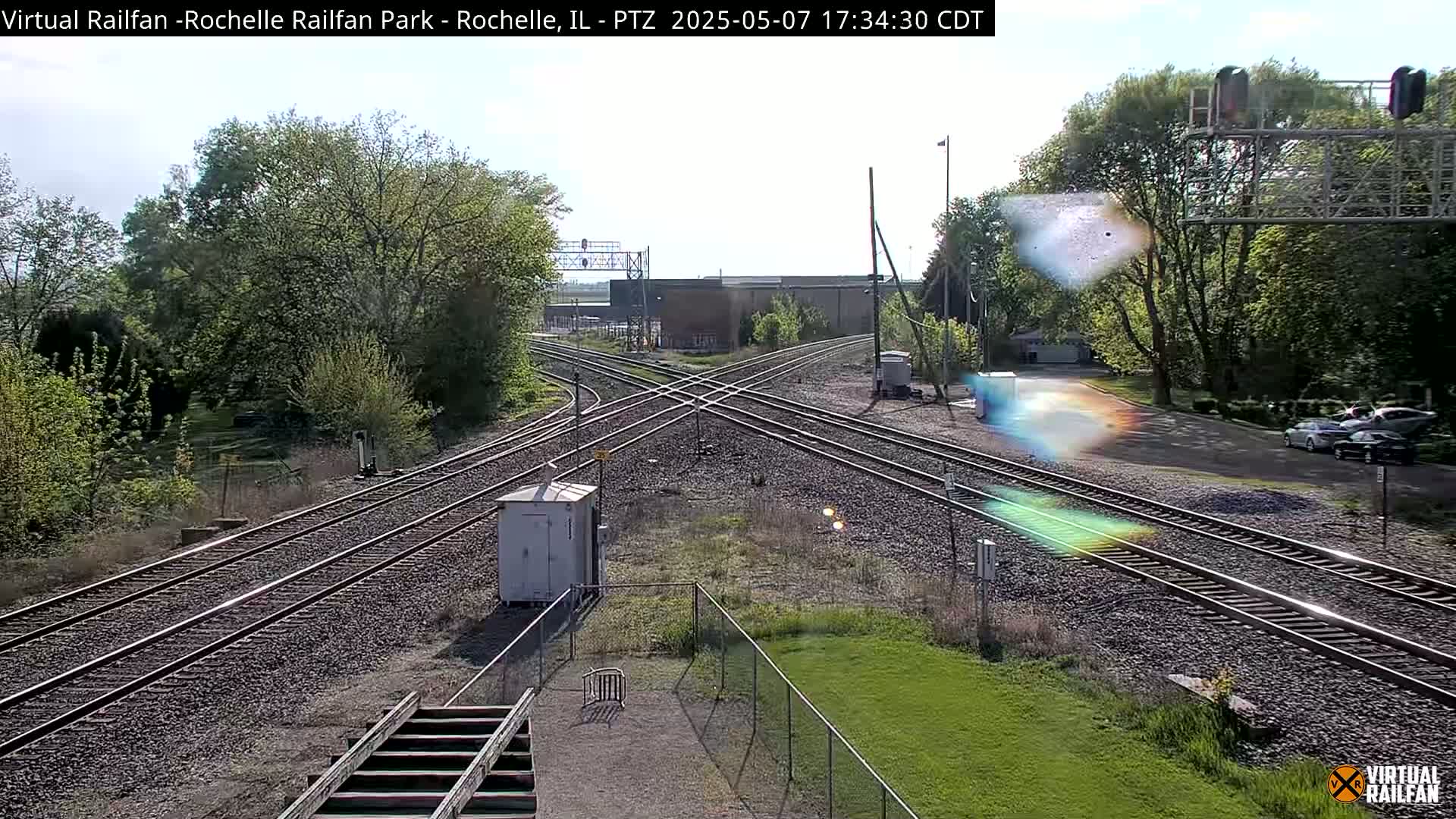 A sunny view of a railroad crossing with multiple tracks, a small building, and trees in the background.