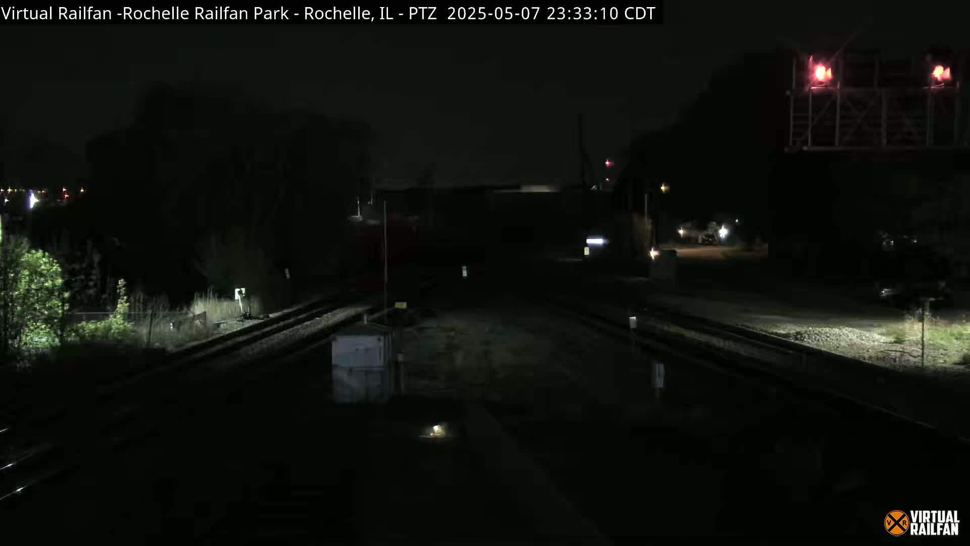 A dark, nighttime view of railroad tracks and a nearby road, with some sparse vegetation visible.