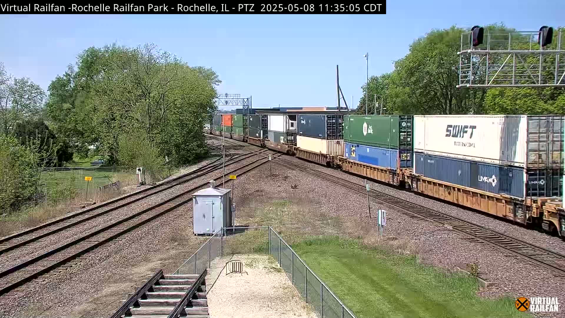 A long freight train carrying numerous shipping containers moves along a railway track under a clear, sunny sky, passing by green trees and a grassy area.