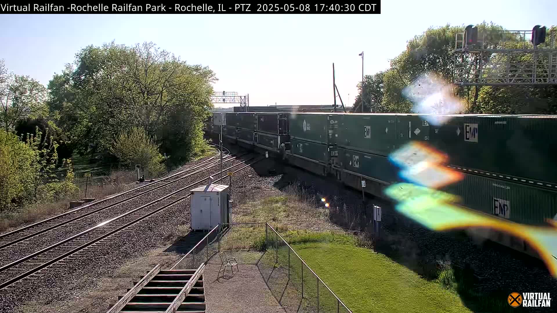 A long freight train carrying multiple green shipping containers passes by a railway under a sunny sky.