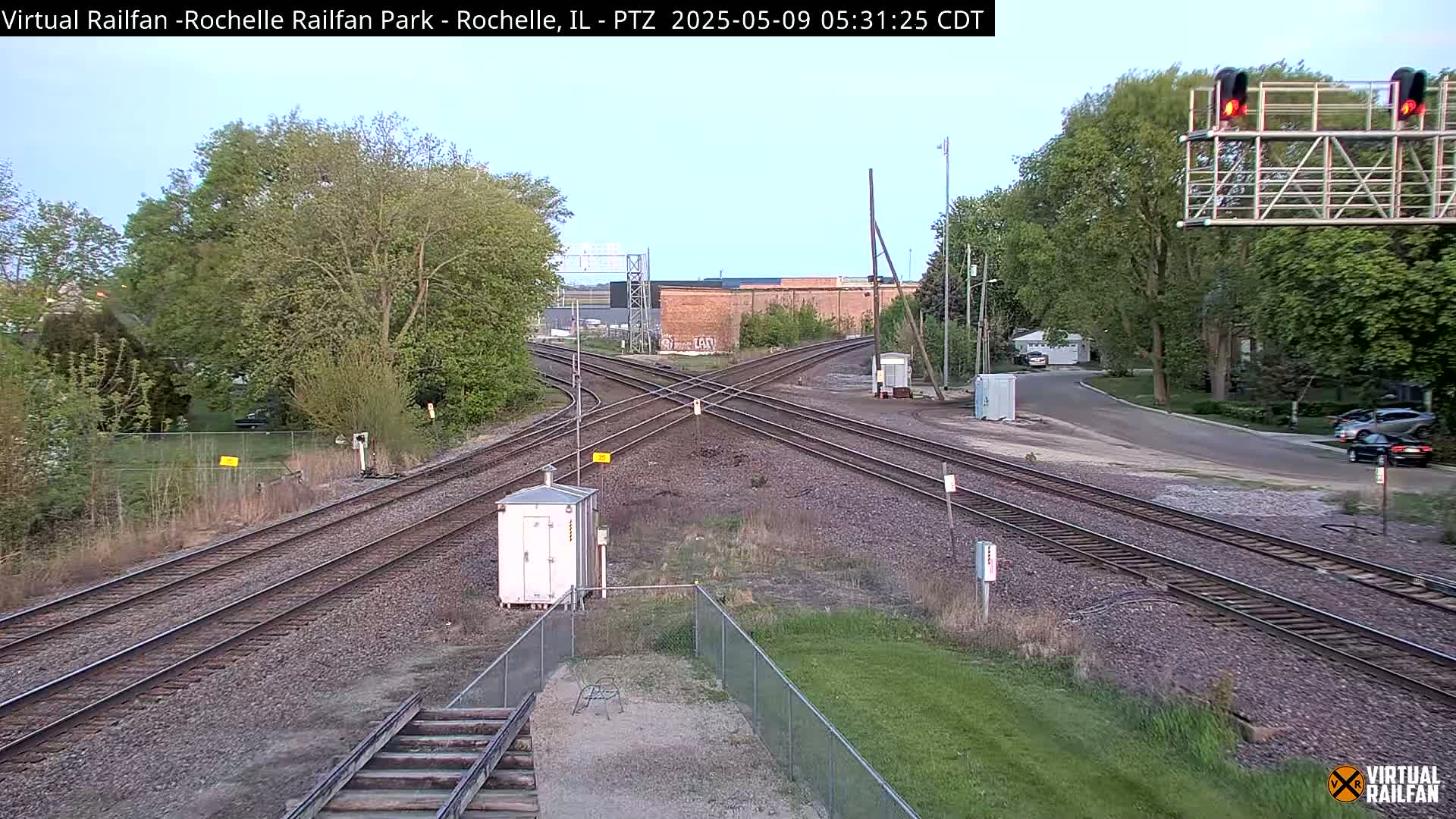 A railroad crossing with several tracks intersecting under a partly cloudy sky, surrounded by trees and a residential area.
