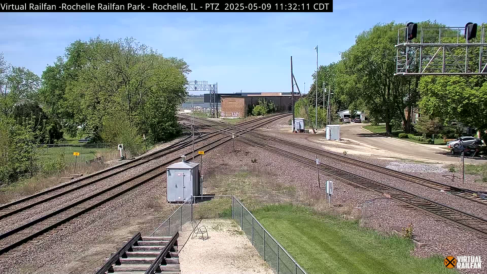 A sunny day reveals a railroad crossing with several tracks, surrounded by green trees and grass, and a small building near the tracks.