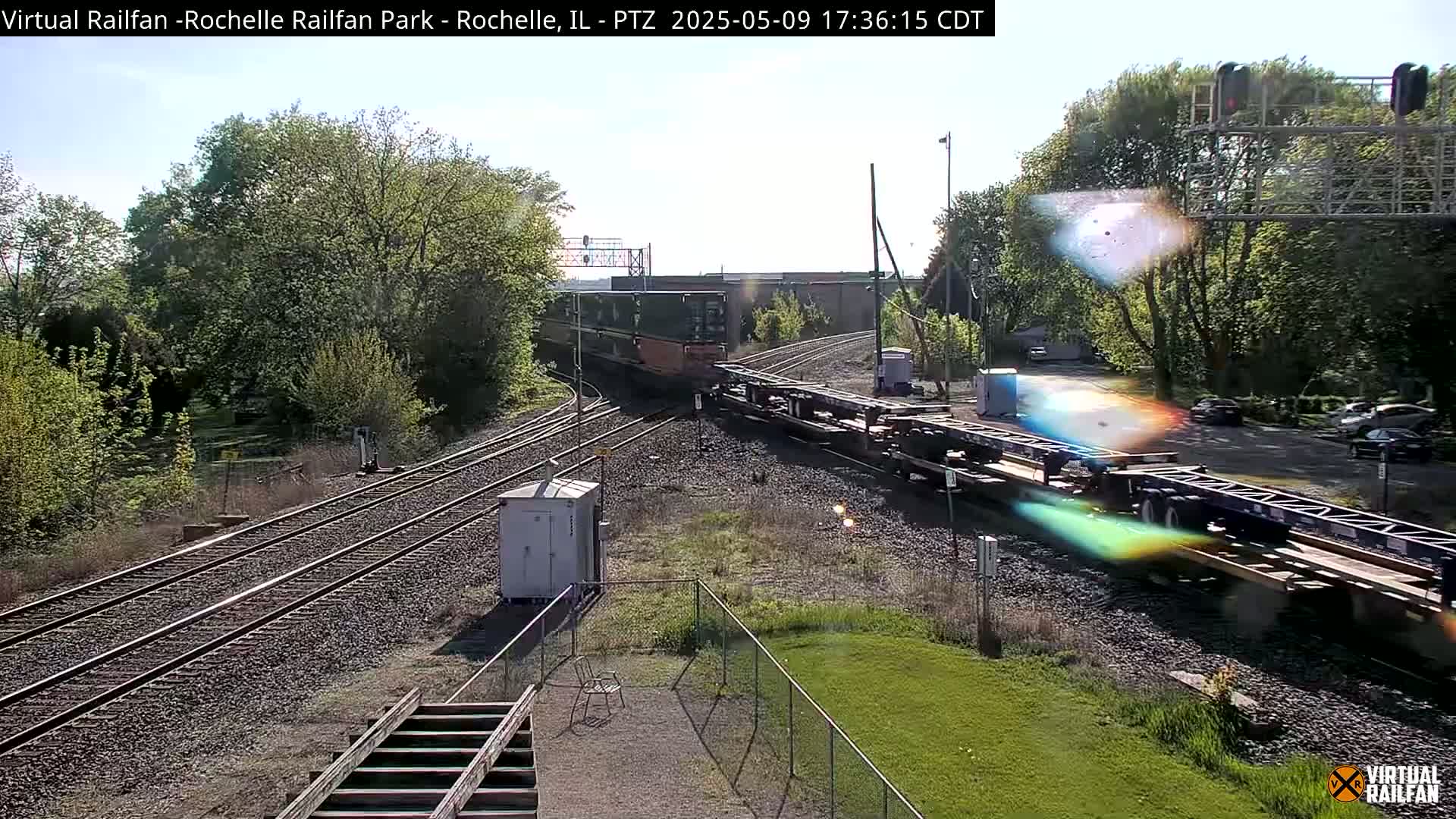 A long freight train passes by on a sunny day, alongside railway tracks that wind through a green, tree-lined area.