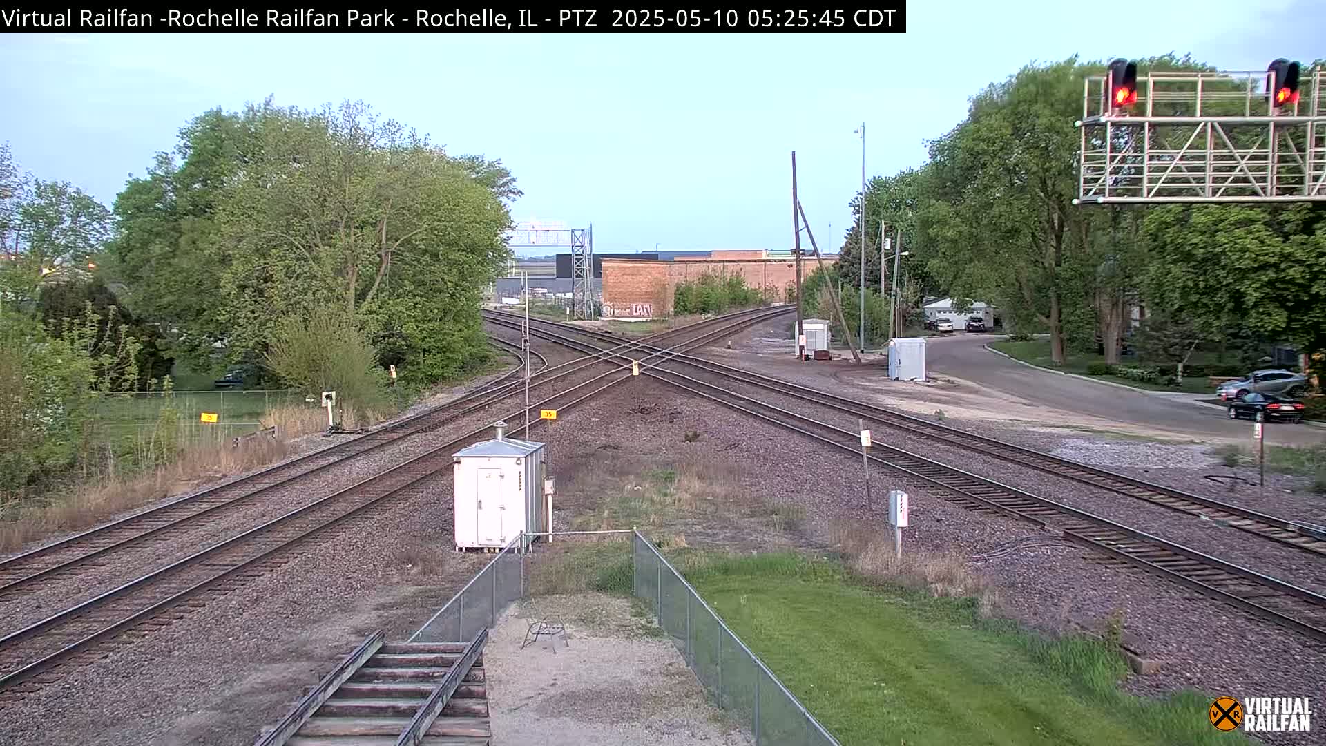 A railroad crossing with several train tracks intersecting under a partly cloudy sky, surrounded by green grass and trees.