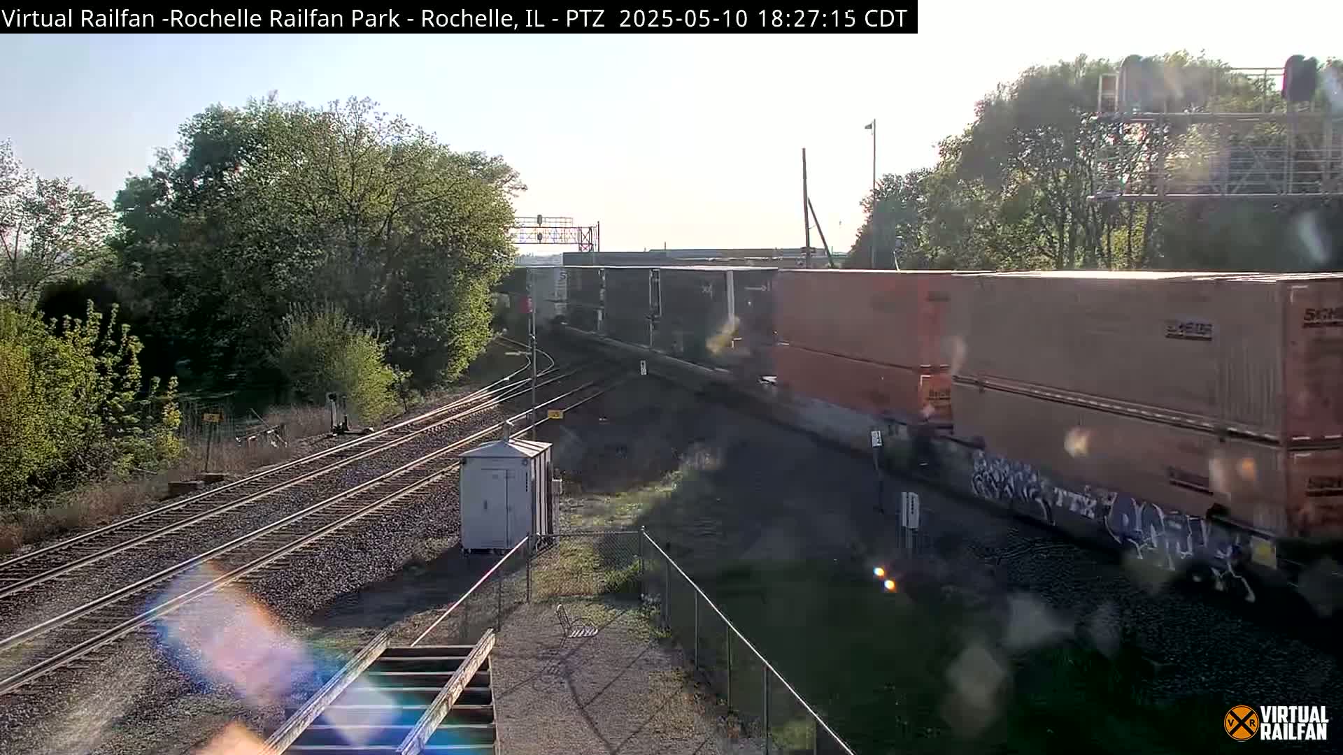 A long freight train with numerous cargo containers passes by railroad tracks on a sunny day.