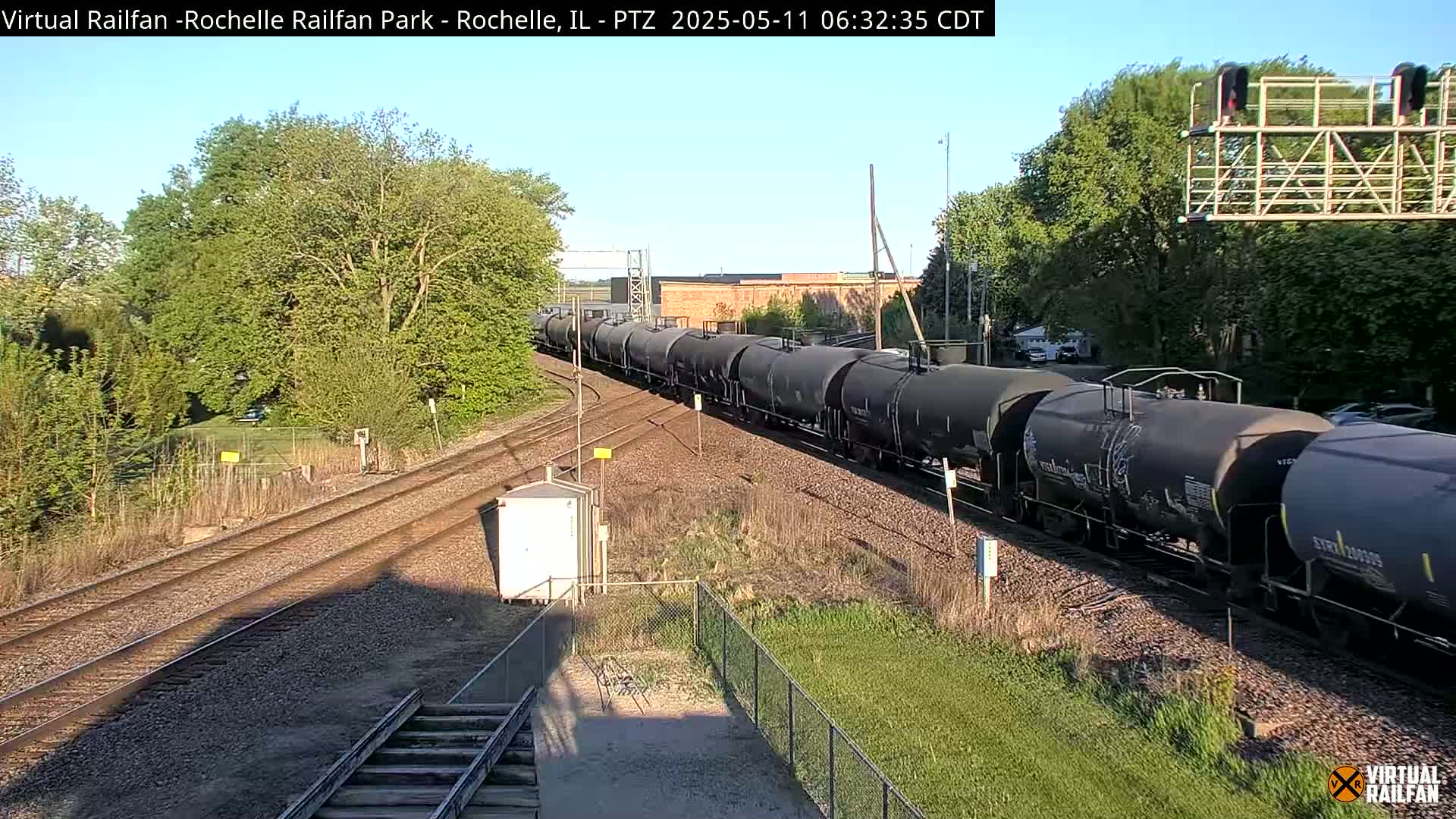 A long freight train of black tanker cars moves along a railway track under a clear, sunny sky, passing through a green landscape with trees and a chain-link fence.