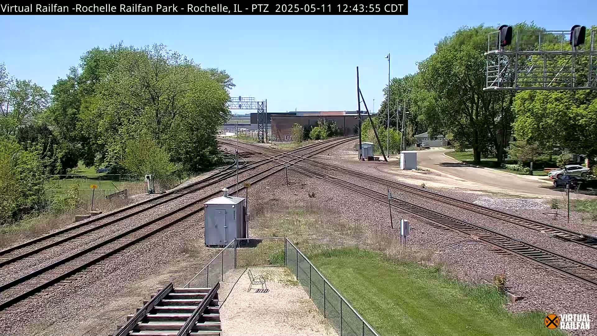 A sunny view of a railroad crossing with multiple train tracks, surrounded by green grass, trees, and a small utility building under a clear sky.