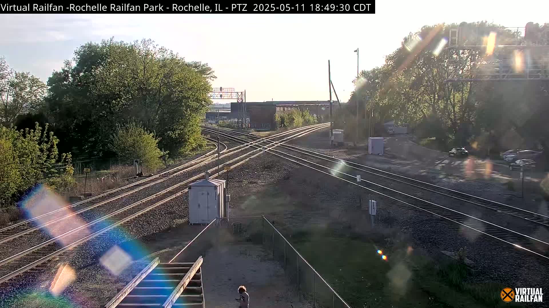 A sunny day at a train crossing with several tracks, surrounded by trees and a chain link fence.