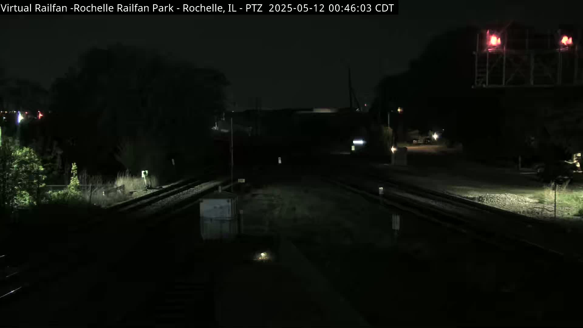 A nighttime view of railroad tracks and a signal light, with dark trees and minimal lighting visible in the surroundings.
