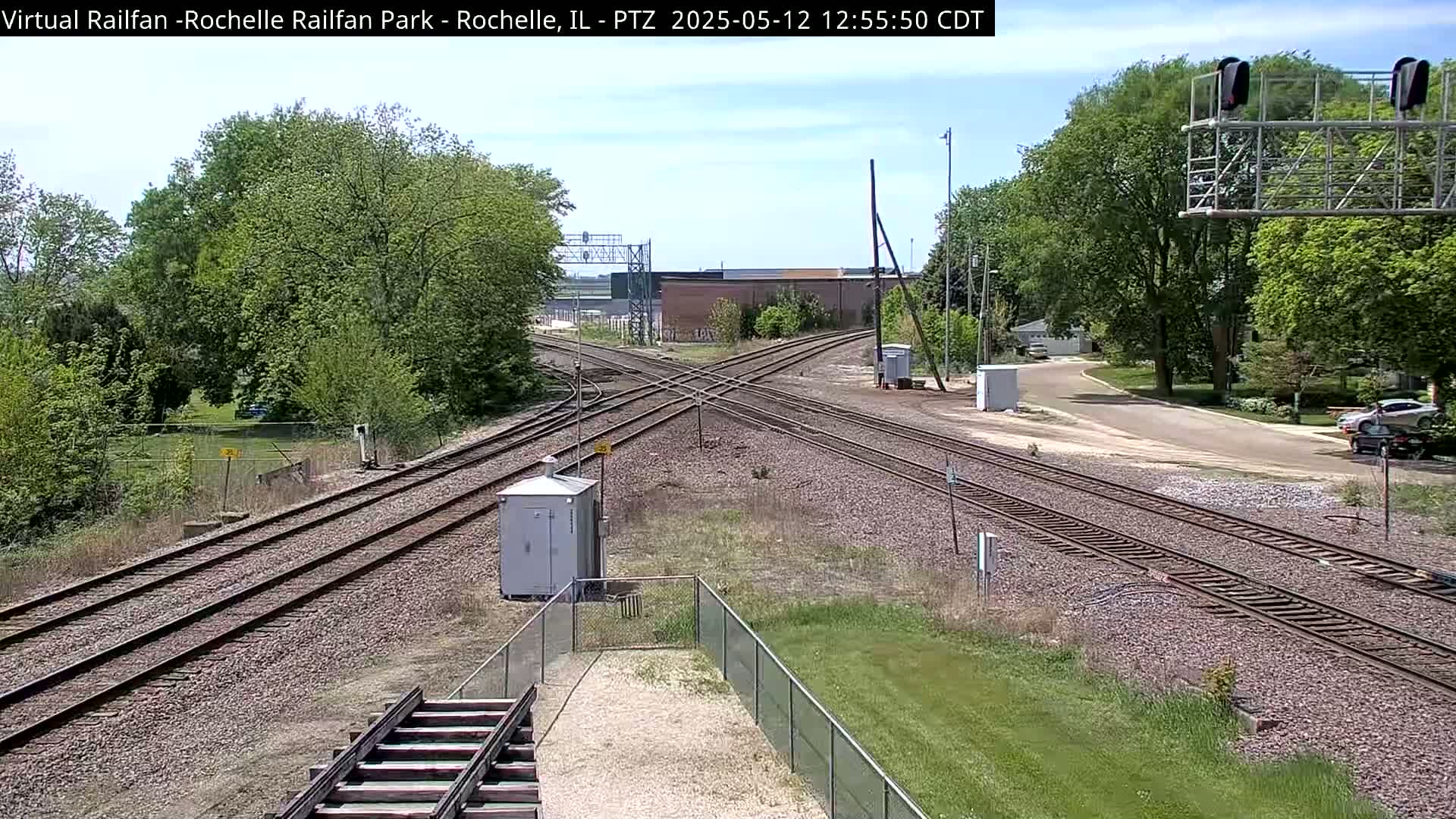 A sunny view of a railroad crossing with multiple tracks intersecting, surrounded by green trees and grass, with a small building and a road visible in the background.