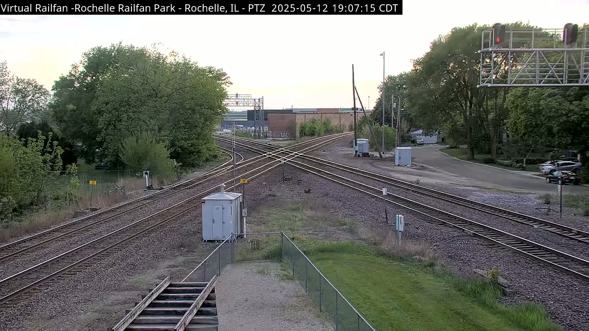 A railroad crossing with multiple tracks intersects a road under a partly cloudy sky, surrounded by green trees and grass.