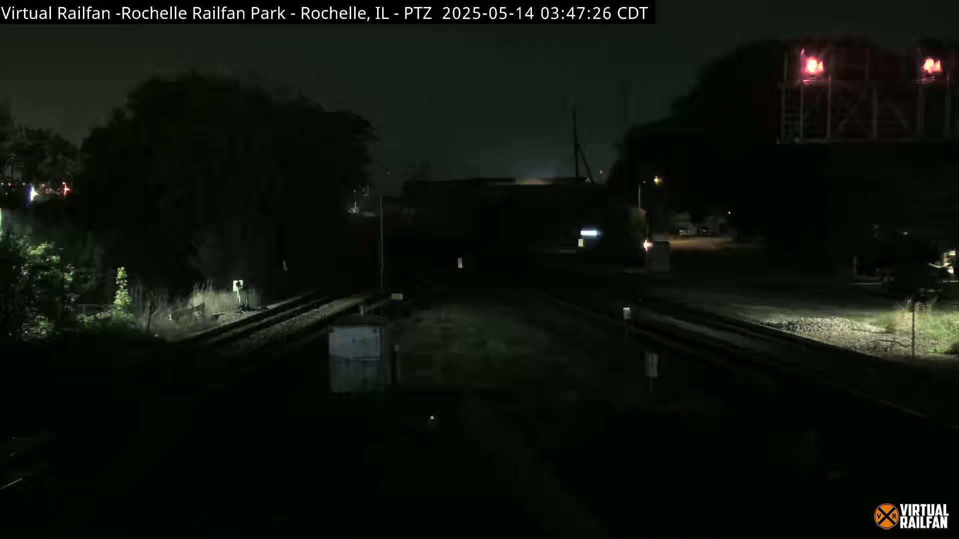 A dark nighttime view of railroad tracks and a nearby structure under a mostly clear sky.