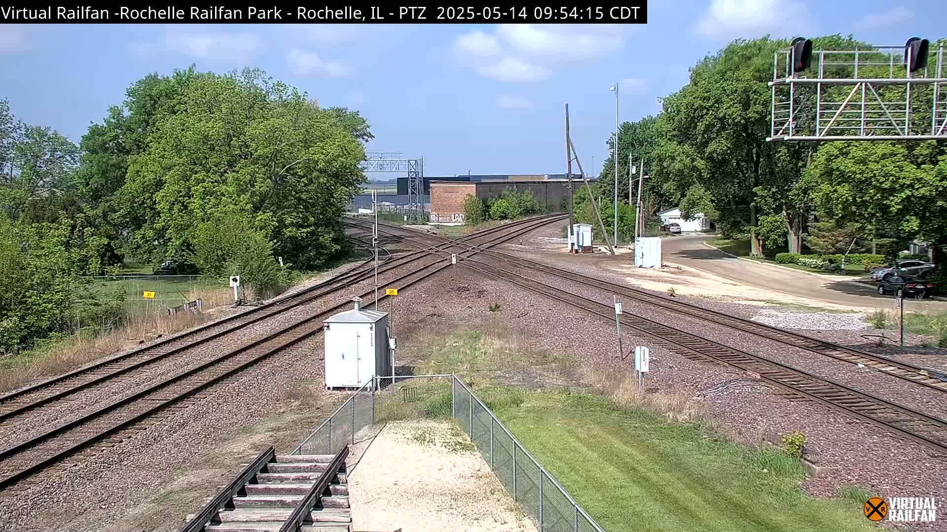 A sunny day at a railroad crossing shows several train tracks intersecting, with green trees and grass surrounding a small, light-colored building near the tracks.