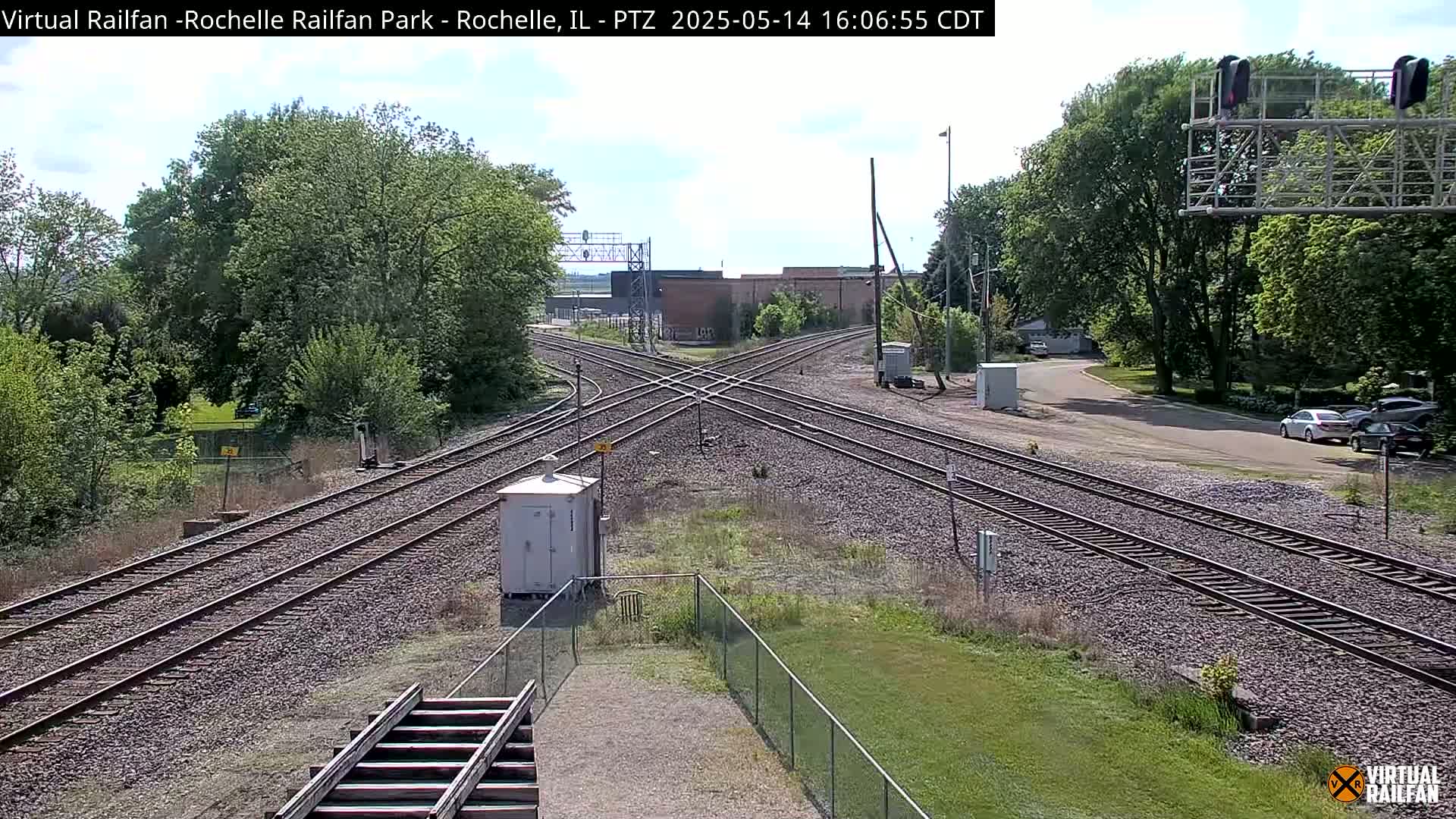 A sunny day shows a railroad crossing with multiple tracks intersecting, surrounded by green trees and grass.