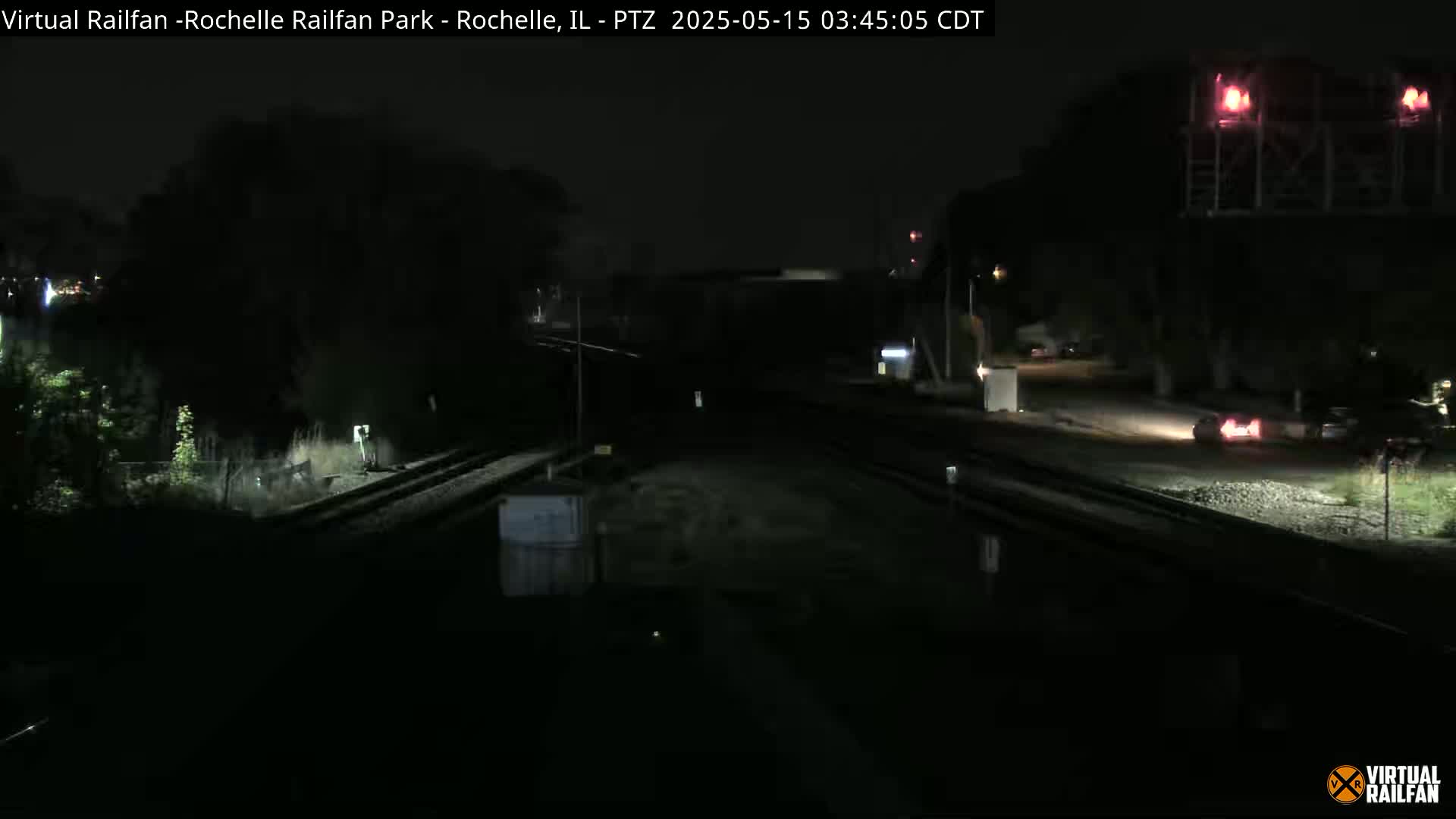 A nighttime view of railroad tracks and a road, with a few vehicles visible in the distance under dark, clear skies.
