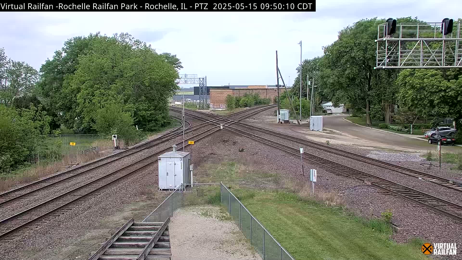 A partly cloudy day shows a railroad crossing with multiple tracks, surrounded by green grass, trees, and a small utility building.