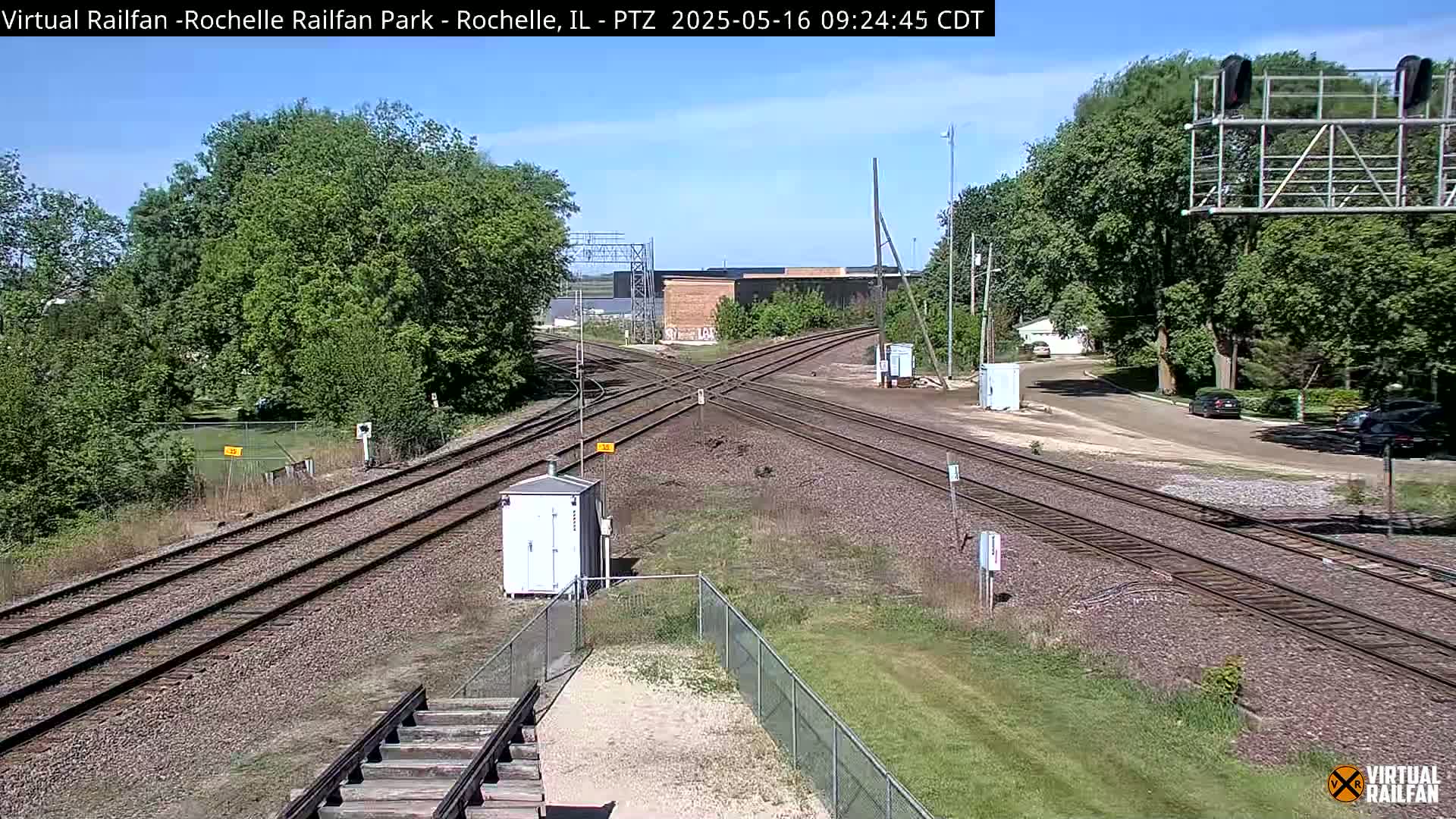 A high-angle view of a sunny railroad crossing with several tracks intersecting, surrounded by green trees and grass.