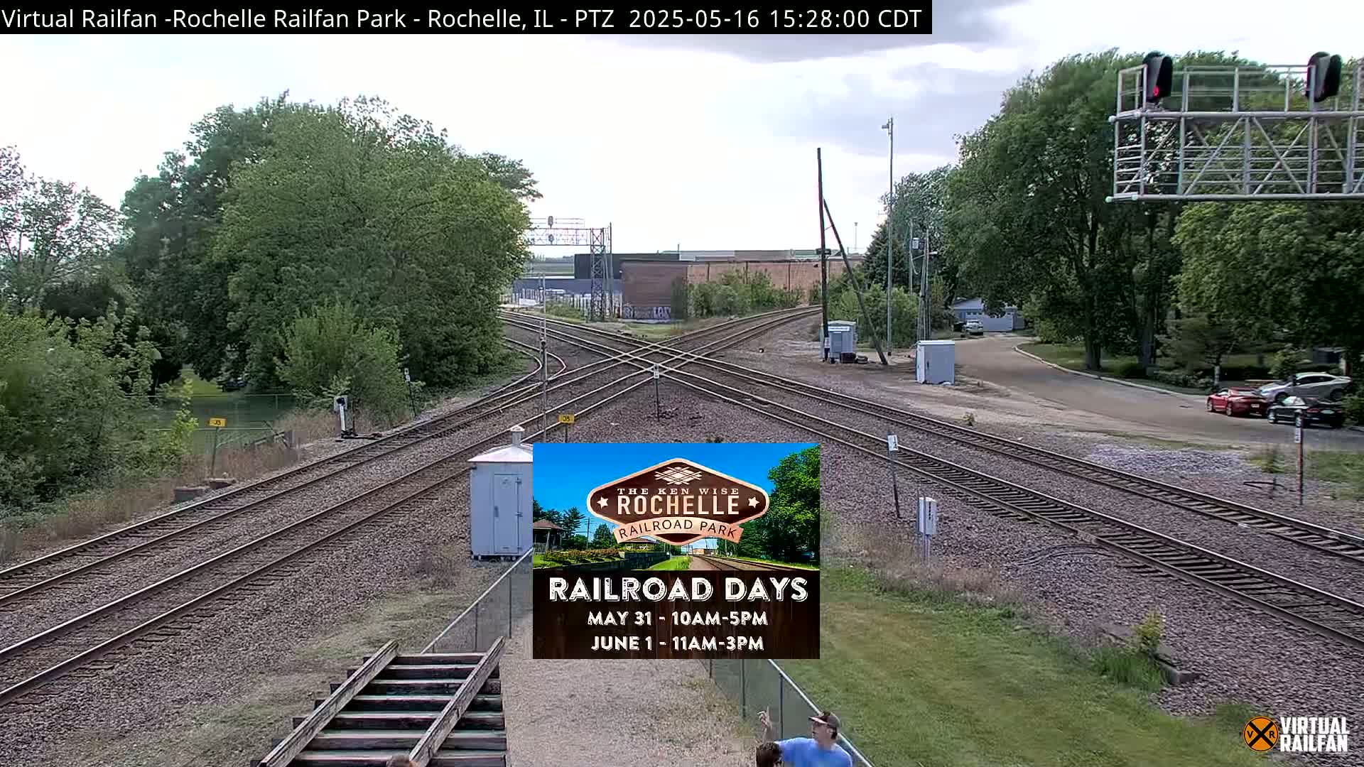 A railroad crossing with multiple tracks is shown on a partly cloudy day, surrounded by green trees and grass, with a few cars parked nearby.