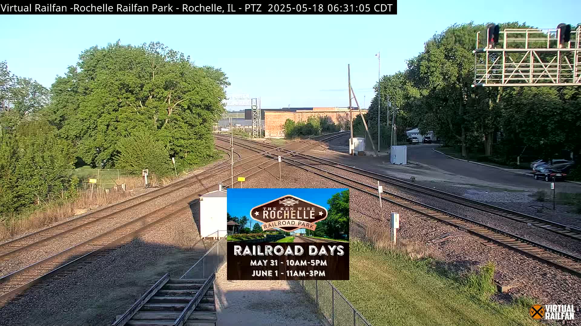 A sunny view of a railroad crossing with multiple tracks, surrounded by lush green trees and vegetation.