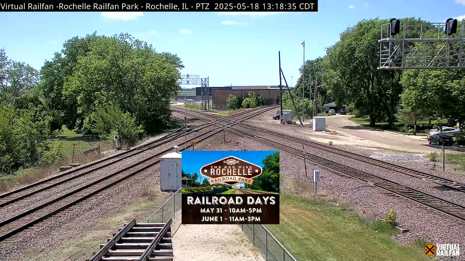 A sunny day reveals a railway intersection with several tracks, bordered by green trees and a paved road, and a small white building near the tracks.