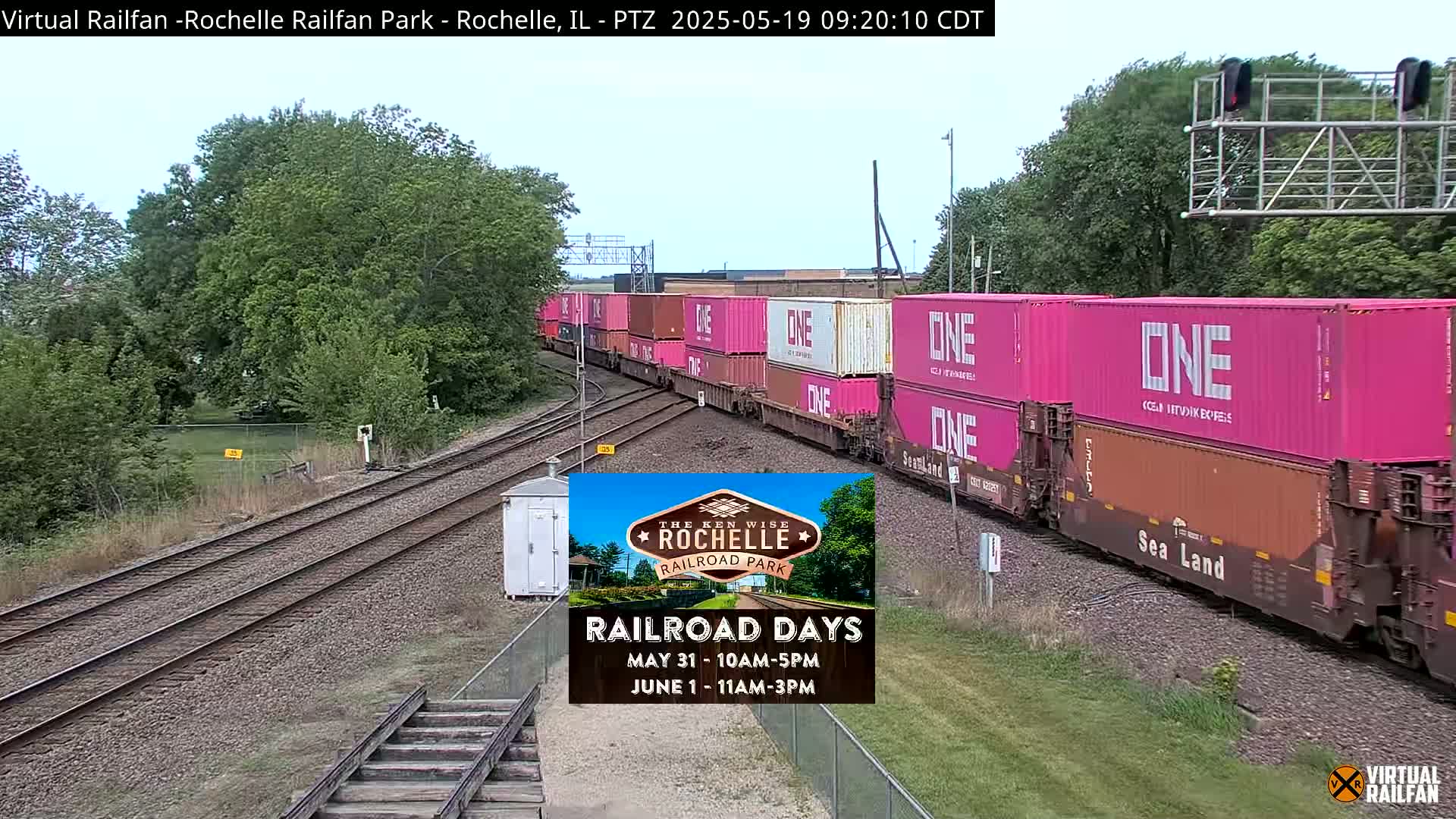 A long freight train carrying bright pink shipping containers moves along railroad tracks under a partly cloudy sky.