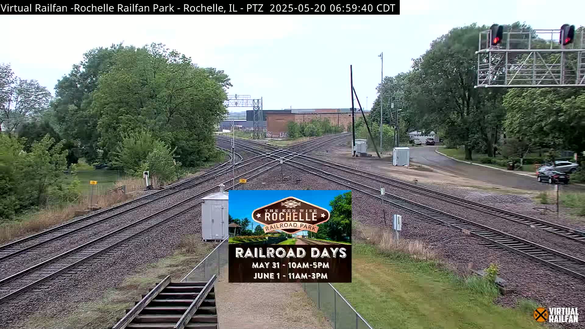 A railroad crossing with multiple tracks and surrounding greenery under an overcast sky.