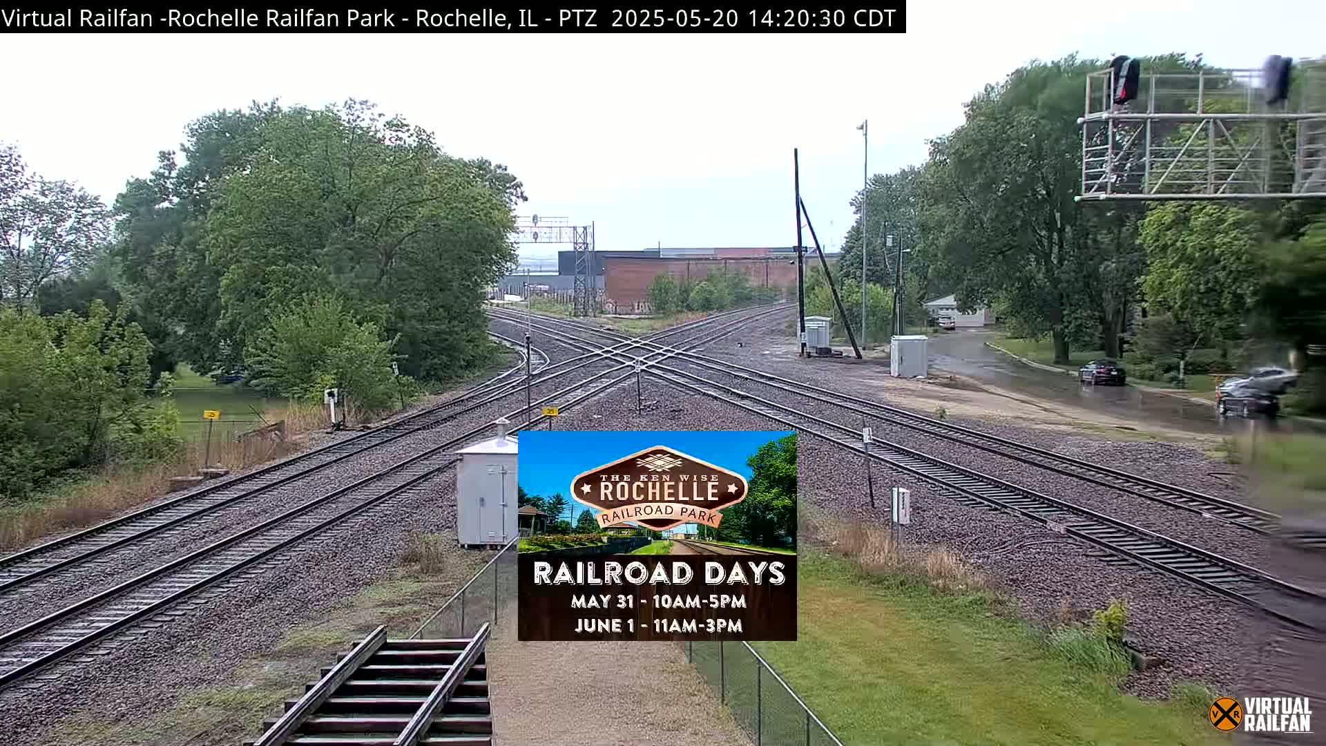 A wet railroad crossing with multiple train tracks intersects a road bordered by lush green trees under an overcast sky.