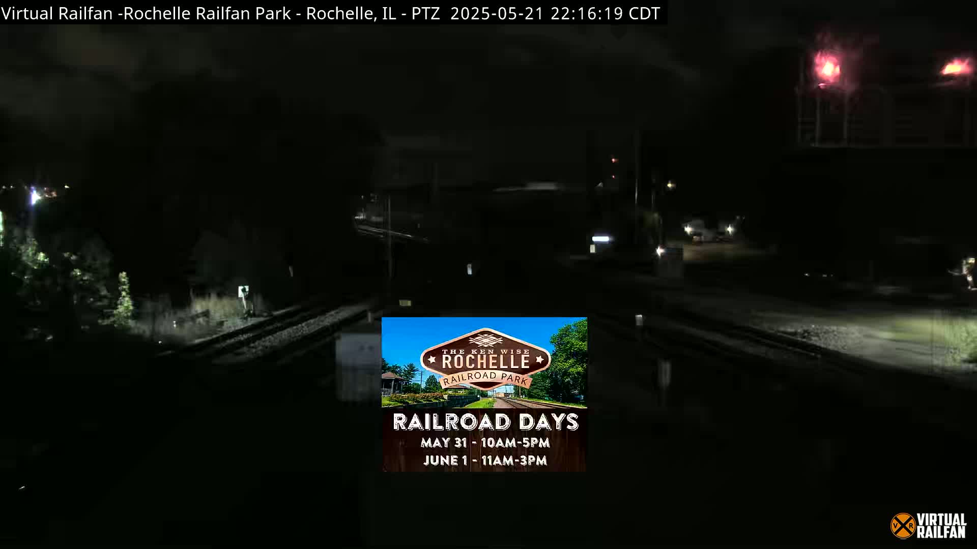 A nighttime, dark view of railroad tracks and surrounding area under a mostly cloudy sky.