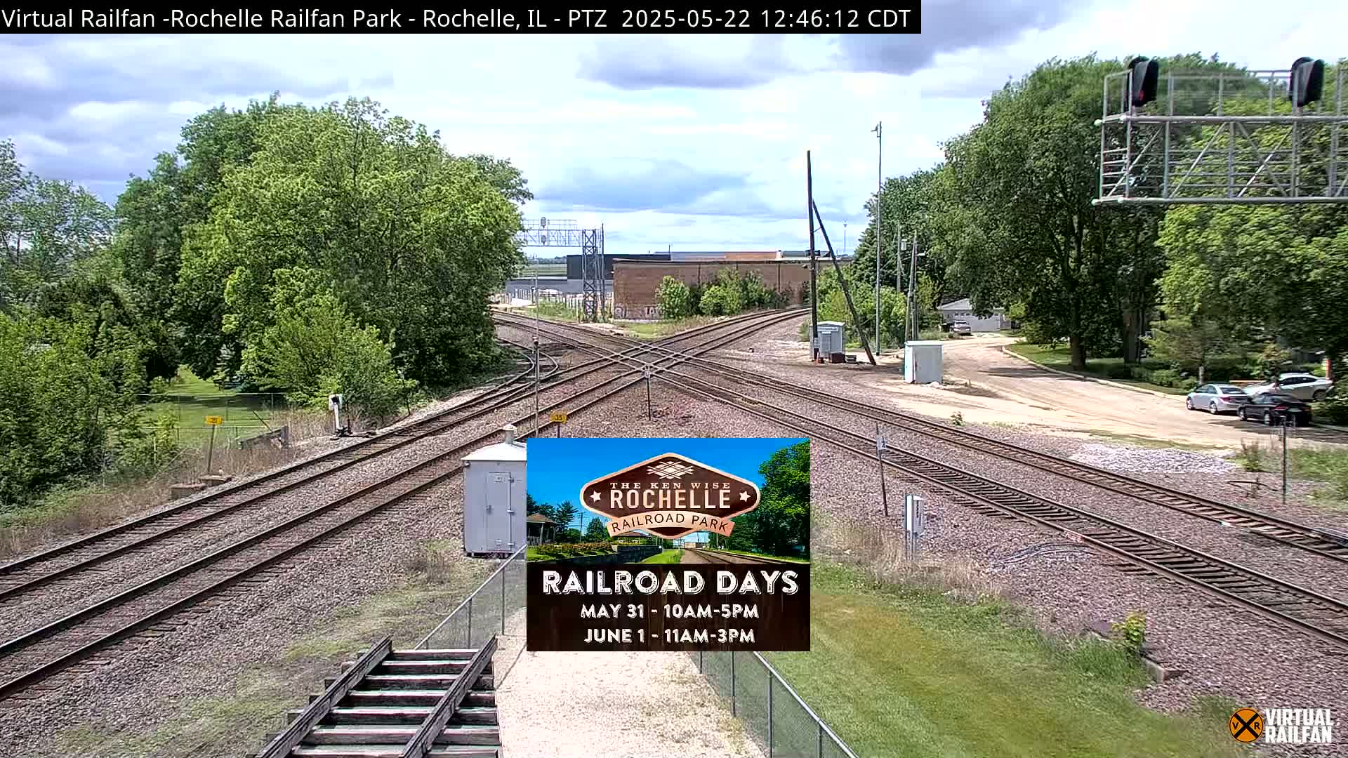 A sunny day at a railroad crossing shows multiple train tracks intersecting, surrounded by green trees and grass, with a few cars visible in the distance.