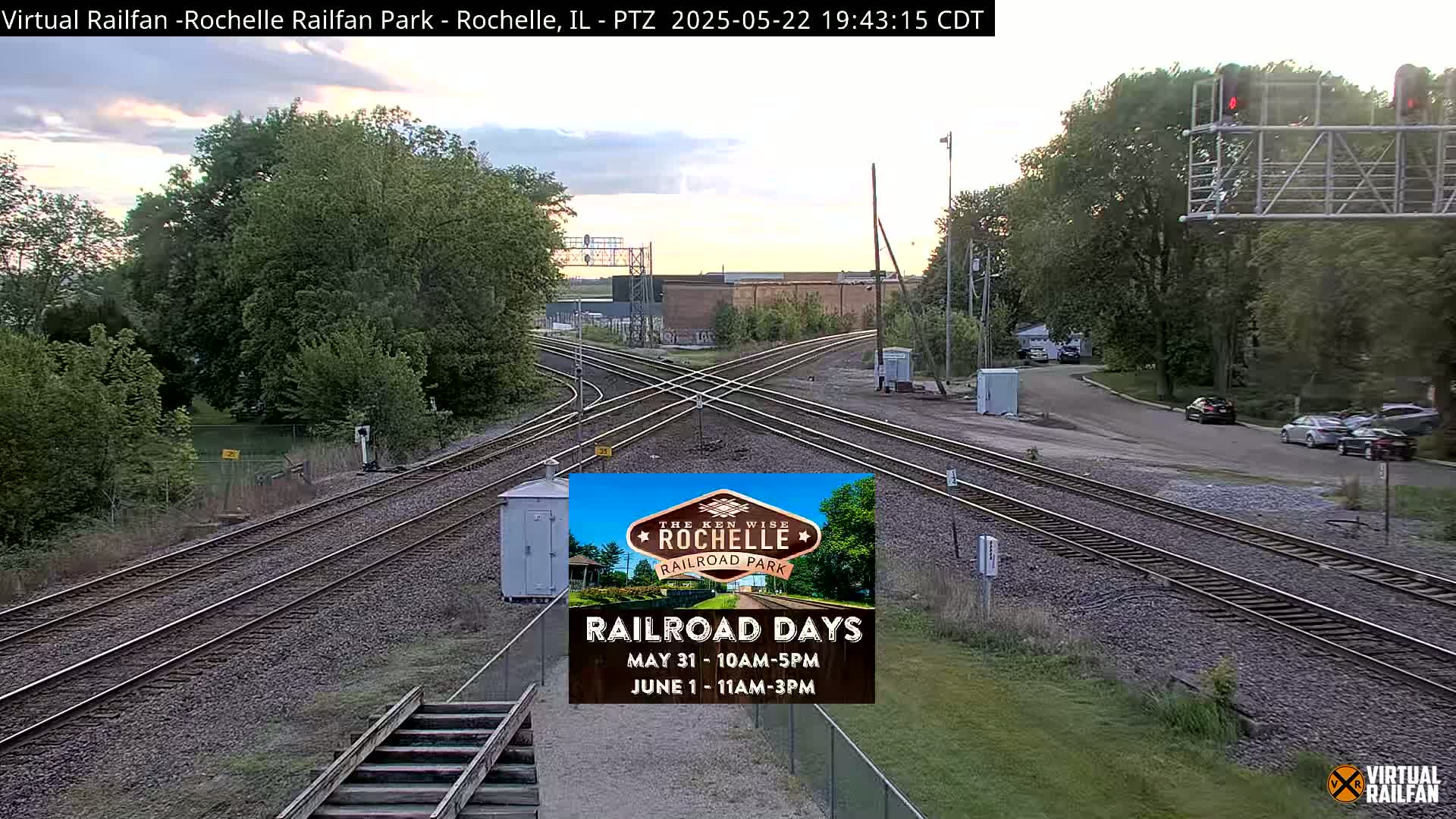 A railroad crossing with multiple train tracks under a partly cloudy evening sky, surrounded by trees and a road with a few parked cars.