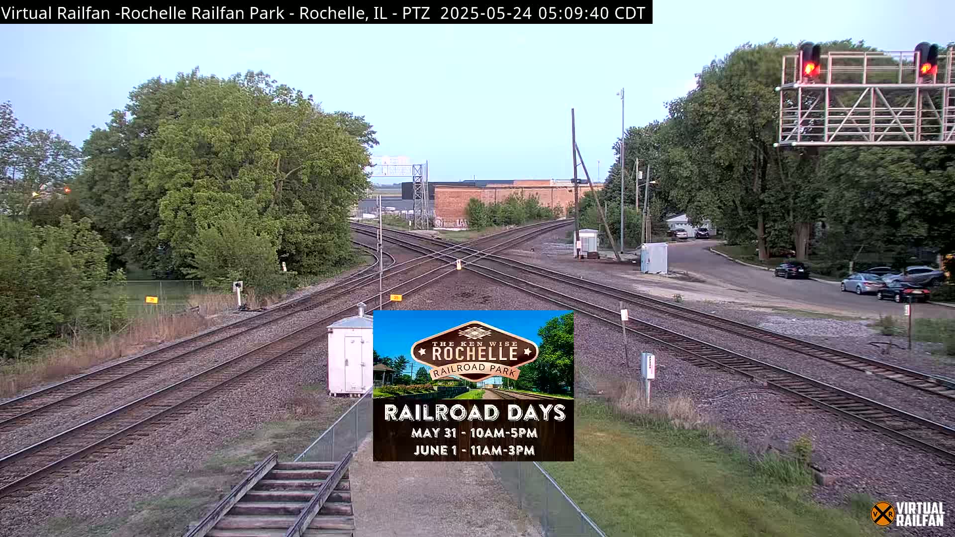 A railroad crossing with multiple tracks and lush green trees under a partly cloudy sky.