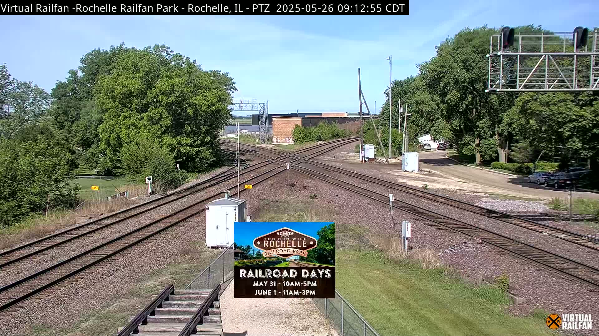 A sunny day at a railroad crossing shows several sets of train tracks intersecting, with green trees and grass surrounding the area.