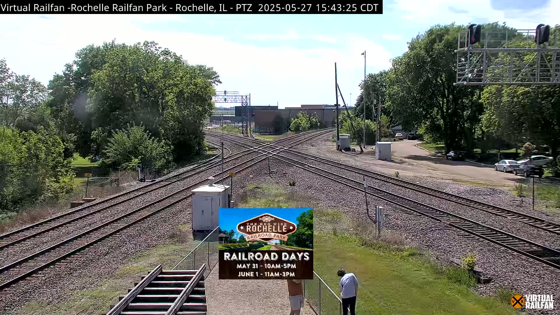 A sunny outdoor view shows a railroad crossing with several train tracks intersecting, surrounded by green trees and grass, and a few cars parked nearby.