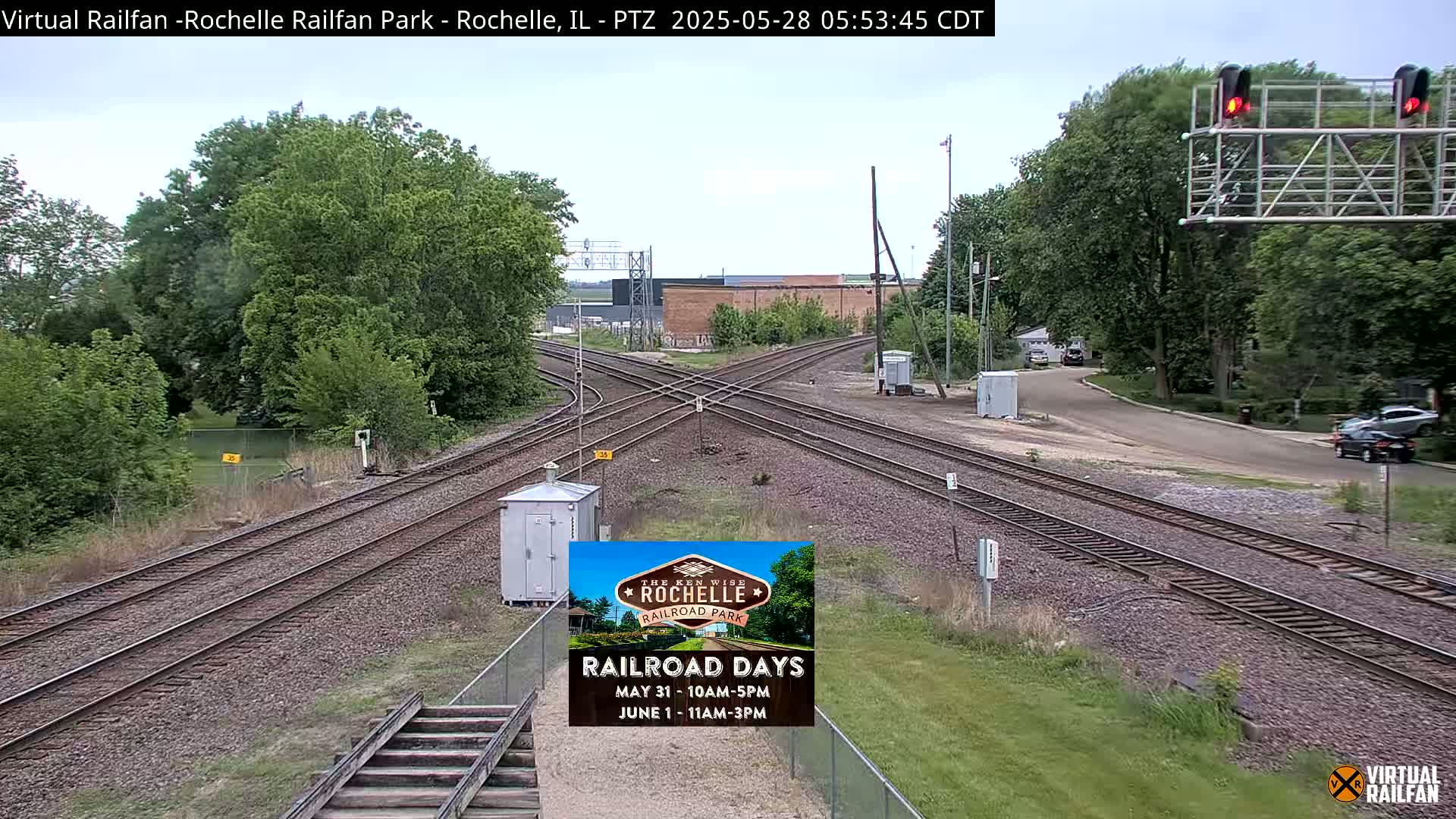A mostly sunny outdoor view of a railroad crossing with several train tracks intersecting, surrounded by green trees and vegetation.