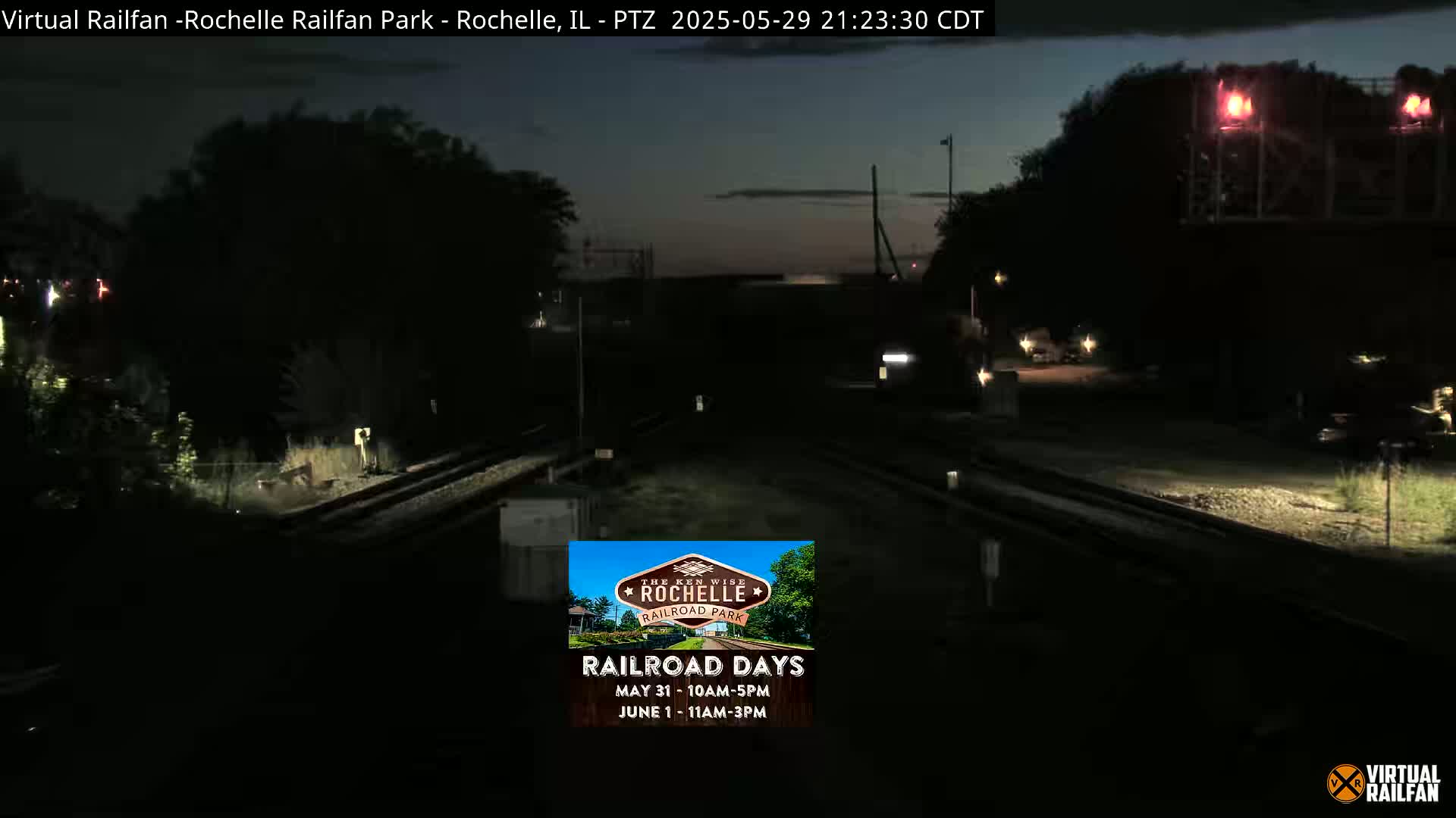 A nighttime view of a dark train yard with railroad tracks and dim lighting under a twilight sky.