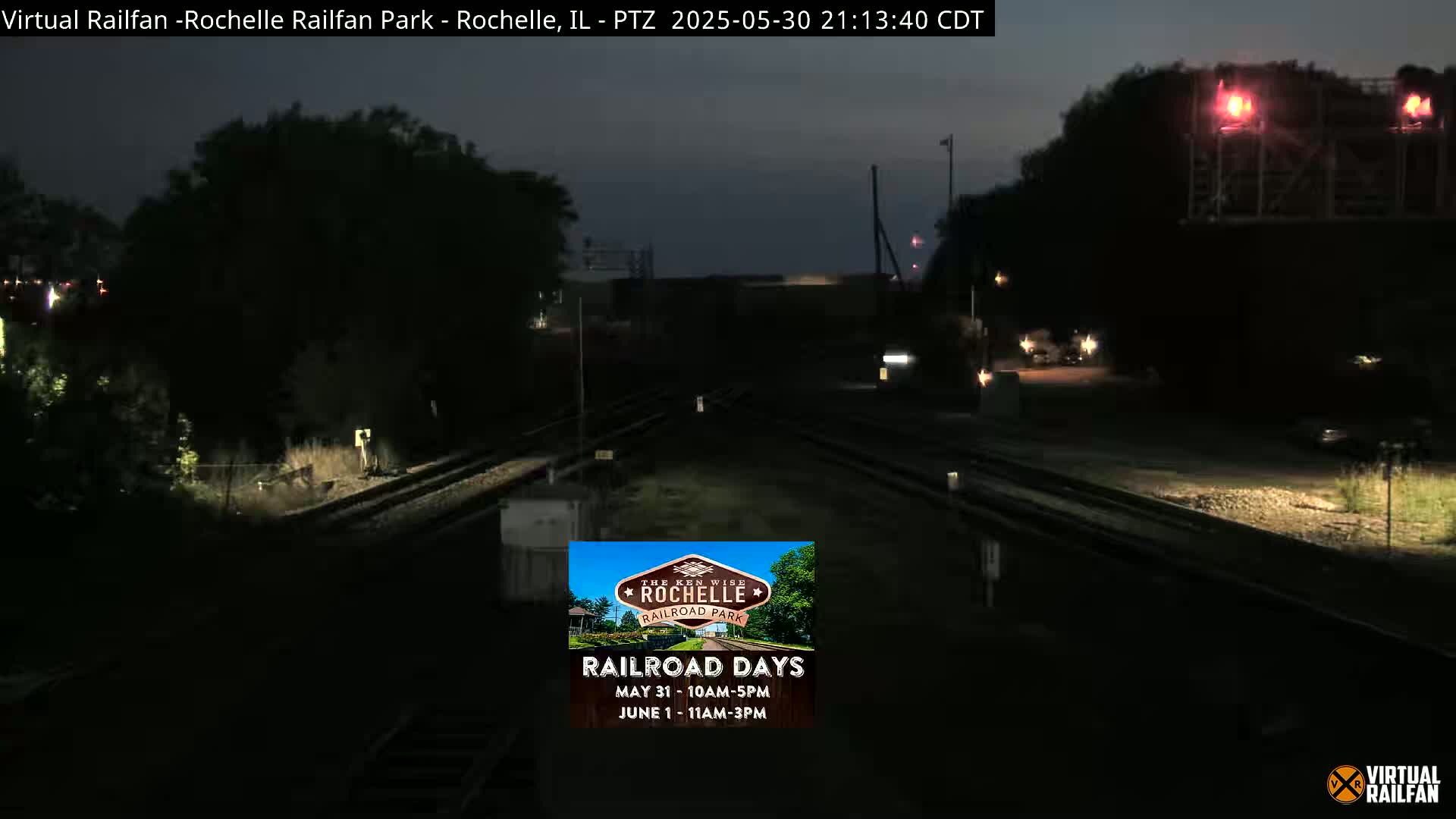 A nighttime view of a railroad track intersection, dimly lit with some distant lights, under a dark sky.