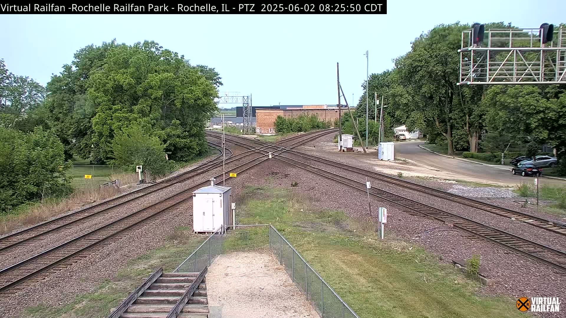 A railroad crossing with multiple train tracks intersects a road under a mostly clear sky, surrounded by green trees and vegetation.
