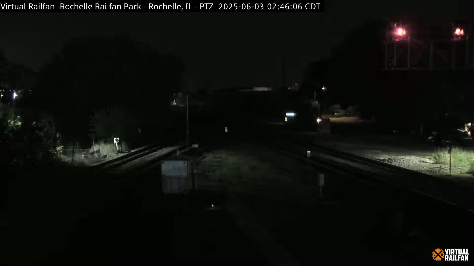 A dark, nighttime view of railroad tracks intersecting, with dimly lit vegetation and structures surrounding the area.