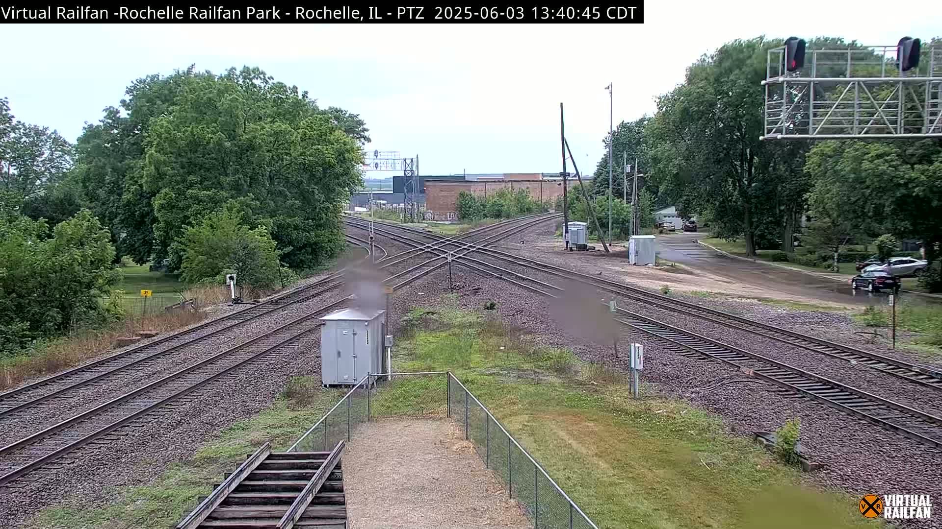 A railroad crossing with multiple tracks surrounded by lush green trees and grass under a partly cloudy sky.