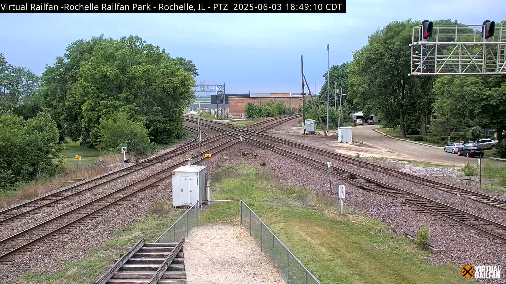 A railroad crossing with several tracks intersects a road, surrounded by lush green trees and grass under a mostly clear sky.