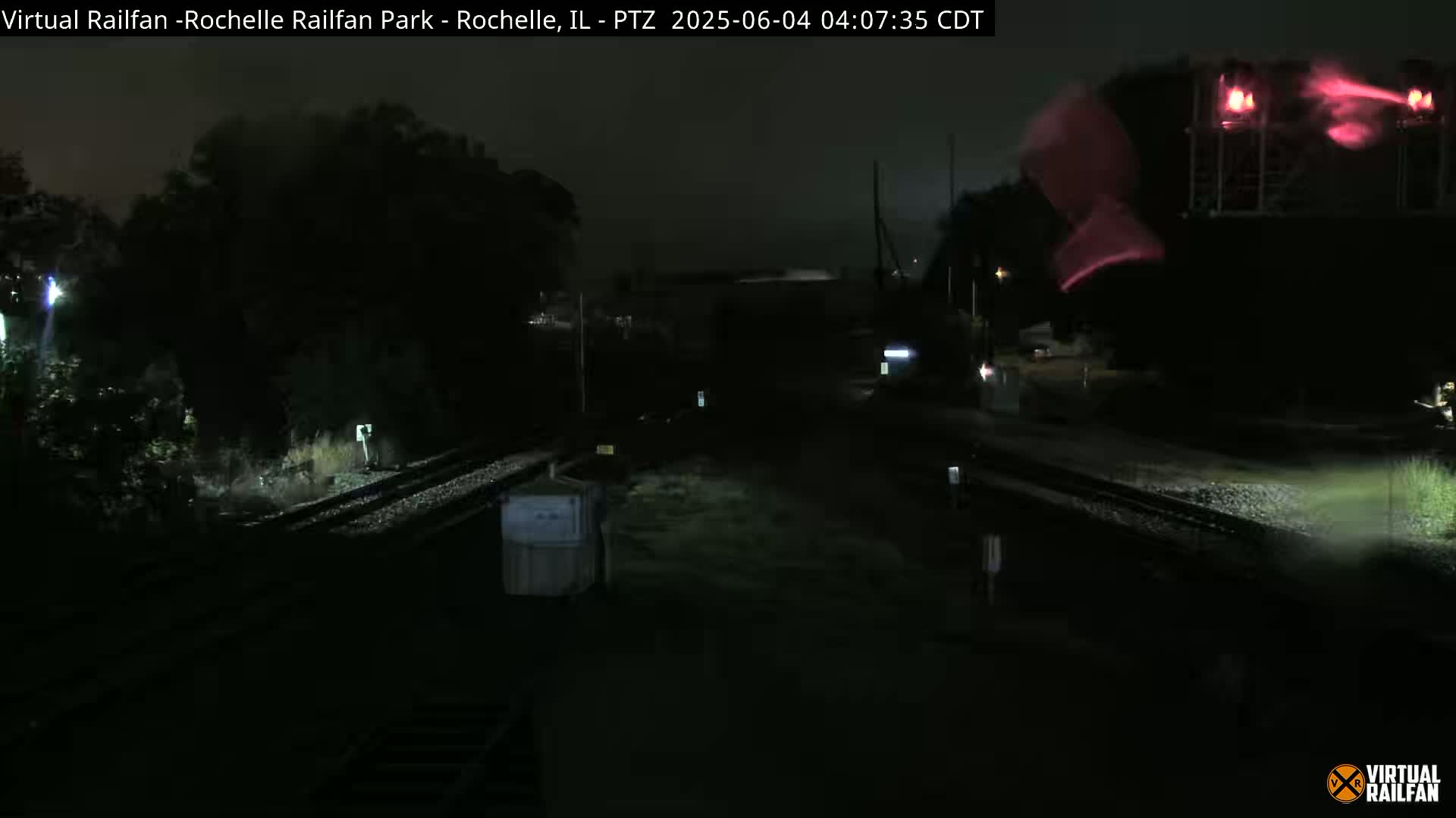 A nighttime view of railroad tracks and a dark structure with red lights, showing wet ground and some vegetation.