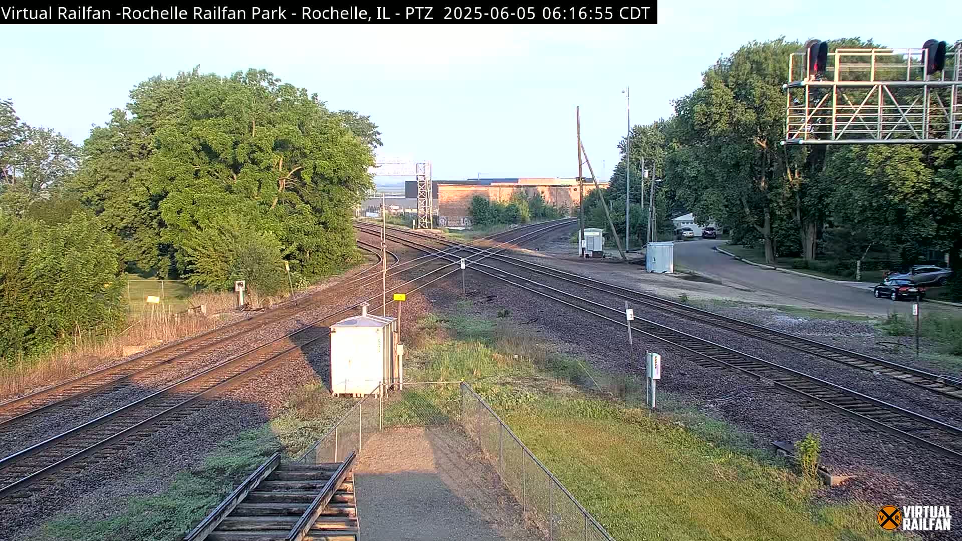 A high-angle, long shot of a railroad crossing under a partly clear sky, with trees lining a road adjacent to the tracks.