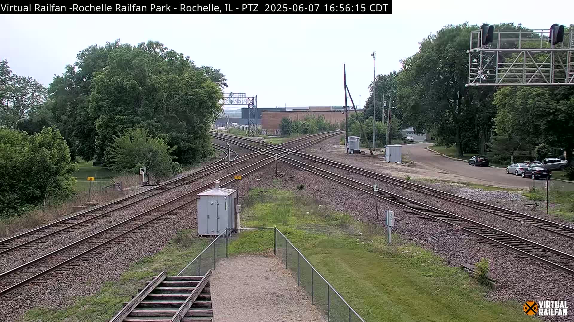 A high-angle view shows a railroad crossing with several train tracks under a partly cloudy sky, surrounded by green trees and grass.