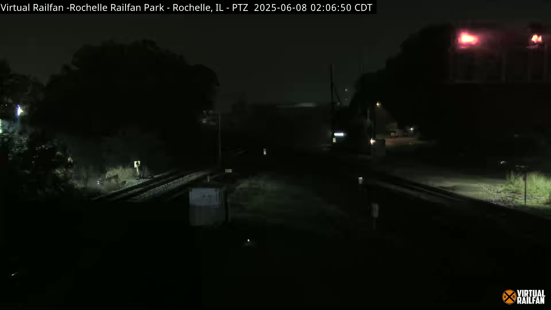 A dark, nighttime view of a railroad crossing with some indistinct structures and vegetation visible in the background.